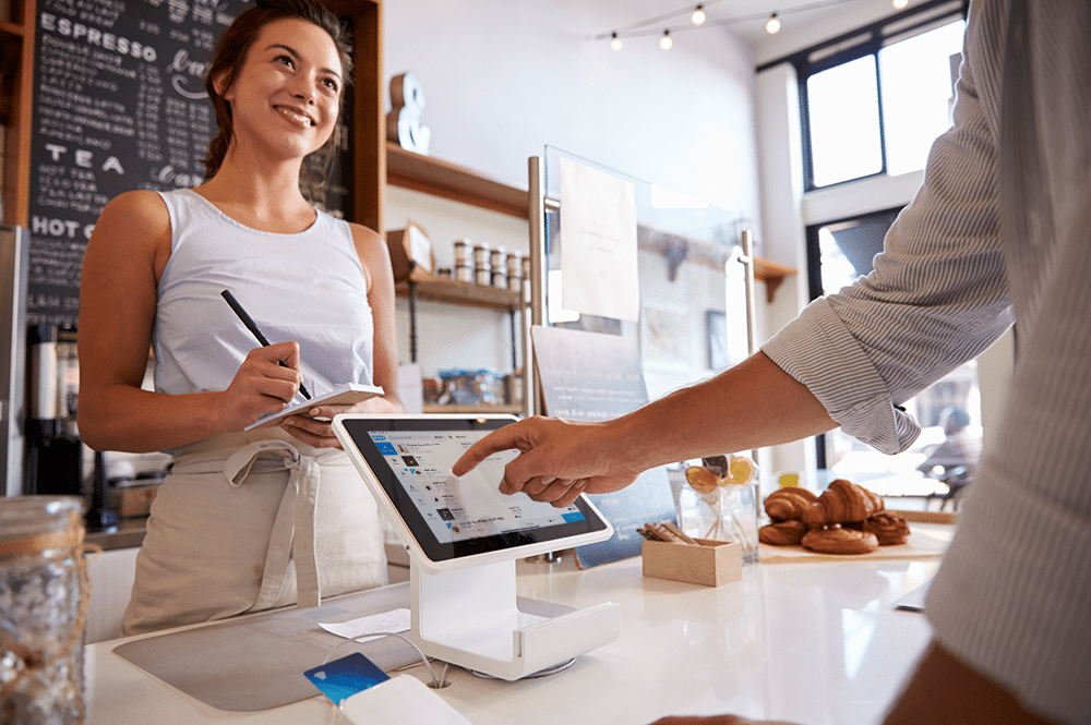 Women Behind A Counter Taking An Order From A Man Selecting An Item From A Square Stand Using Foosales Pos.